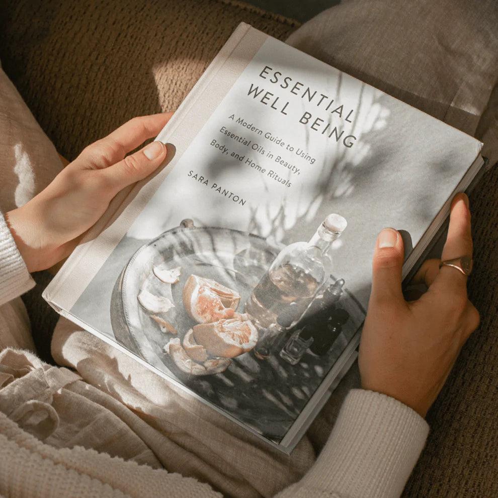 Hands holding "Essential Well Being" book with a cover image of essential oils and citrus.
