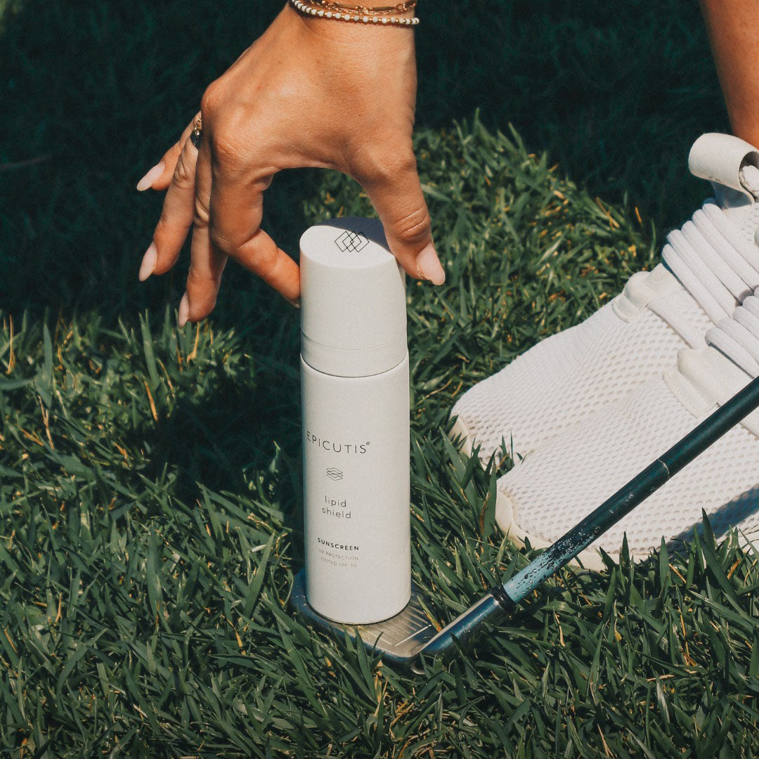 Hand holding a white Epicutis Lipid Shield SPF 30 sunscreen bottle on green grass next to a golf club and white shoe.