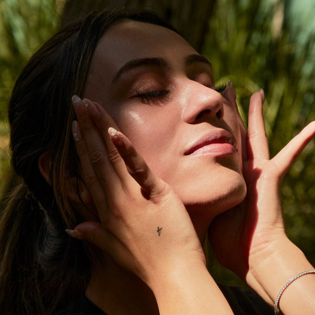 Woman with closed eyes applying Lipid Shield SPF 30 sunscreen, glowing in sunlight, surrounded by green foliage.