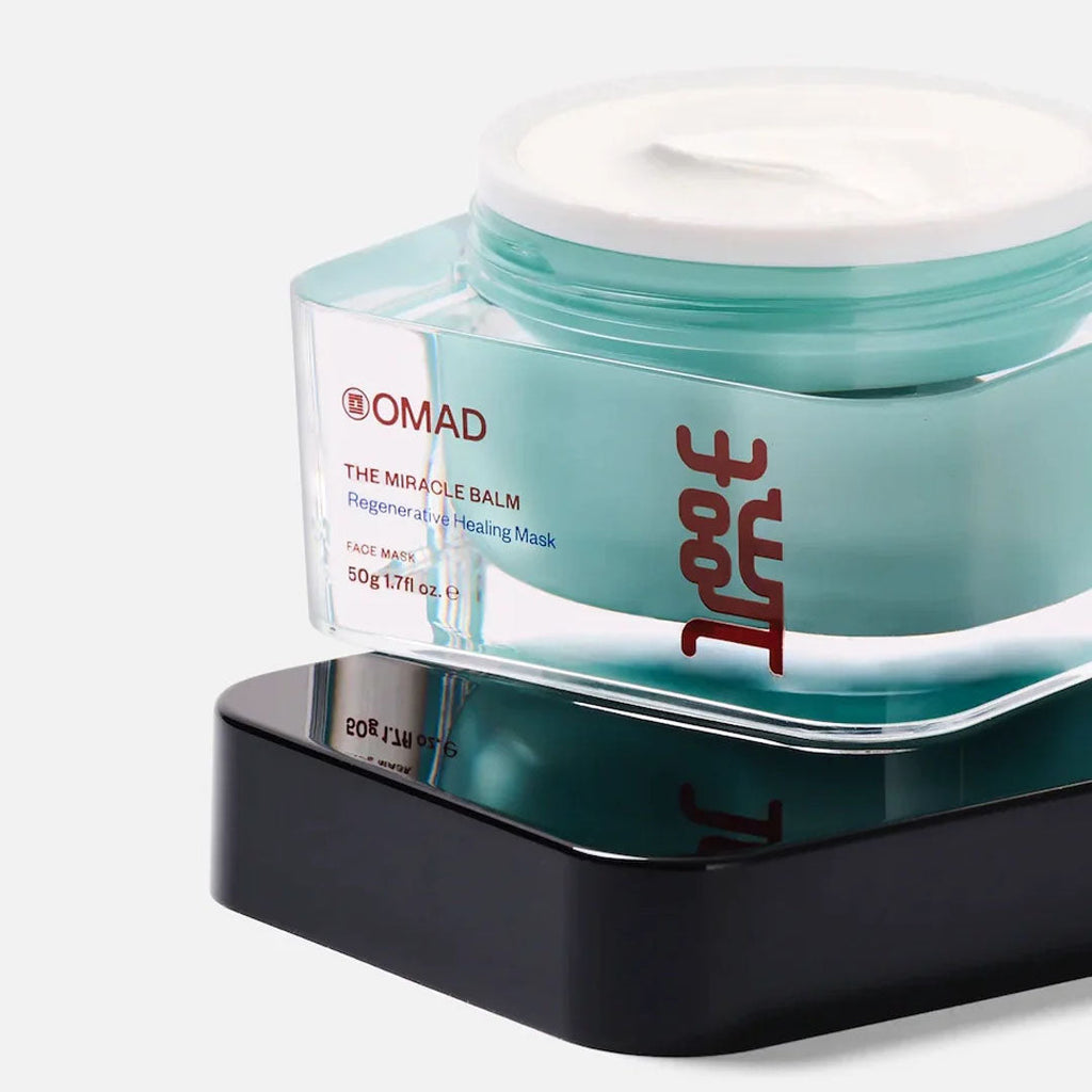 OMAD Miracle Balm Regenerative Healing Mask in a clear teal jar with white cream, on a black base.