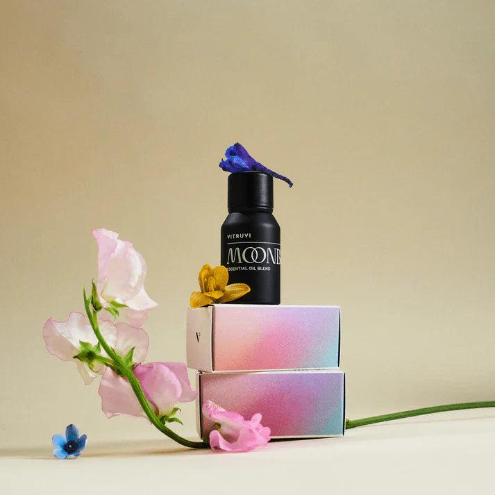 Black Vitruvi Moonbeam essential oil bottle on iridescent boxes with pink and blue flowers.