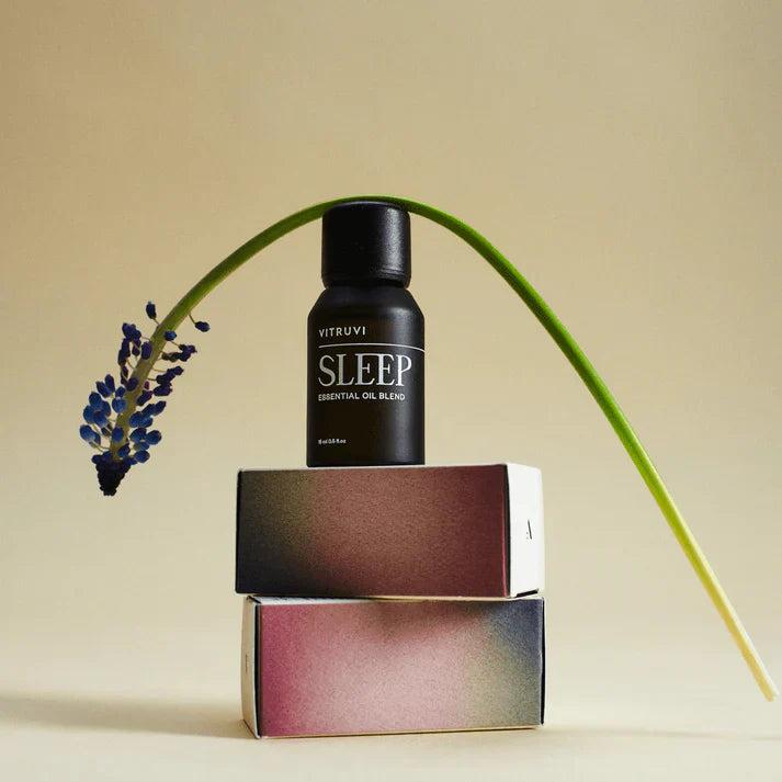 Black Vitruvi Sleep essential oil bottle on two gradient boxes with a curved green stem and purple flower.
