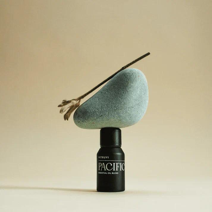 Vitruvi Pacific Essential Oil Blend in a black bottle, balancing a smooth grey-blue stone and dried botanical.